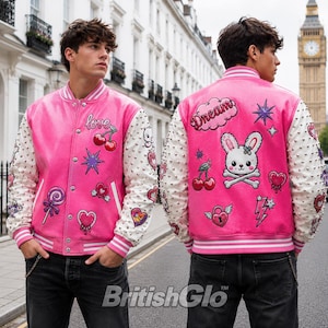 Puede incluir: Una chaqueta universitaria rosa brillante con mangas blancas con tachuelas plateadas. El frente muestra parches de cerezas, una piruleta y la palabra love. La parte posterior presenta un gran parche de calavera de conejo, la palabra Dream e iconos.