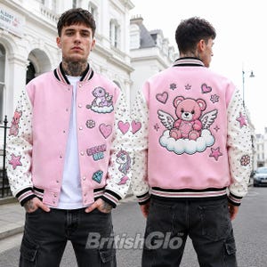Op de afbeelding: Een roze varsity-jack met witte mouwen voorzien van zilveren studs. De voorkant toont patches van een draak, een eenhoorn, een diamant en de tekst "DREAM BIG". De achterkant heeft een grote geborduurde patch van een gevleugelde teddybeer op een wolk.