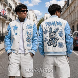 Op de afbeelding: Een wit en lichtblauw varsity-jack met geborduurde patches van een konijn, sterren en vlammen, met de tekst "DREAM BIG," "NO RULES," en "STAY WEIRD." De lichtblauwe mouwen zijn voorzien van zilveren studs en gestreepte boorden.