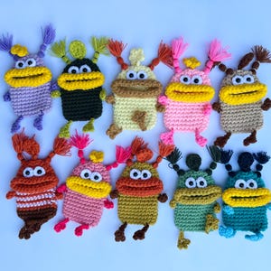 Op de afbeelding: Een verzameling van elf handgemaakte gehaakte monsterpoppen in verschillende kleuren. Elke pop heeft een grote gele mond, twee ogen en twee antennes met garenkwastjes. De poppen zijn in tinten paars, zwart, beige, roze, bruin, oranje, groen en blauw.