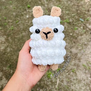 May include: White crocheted llama amigurumi toy with brown ears and black eyes. The toy is being held in a hand. The text "PDF PATTERN" is visible in the lower right corner of the image.