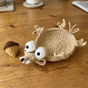 Puede incluir: Un personaje de ganchillo hecho a mano en beige con ojos blancos grandes y una nariz negra, tumbado boca arriba. Junto a él hay una pequeña bellota con gorro marrón y cuerpo amarillo. El personaje tiene un hocico largo y dientes blancos pequeños.