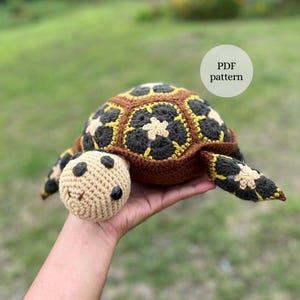 Op de afbeelding: Een gehaakte schildpad met een bruin hoofd, zwarte ogen en een lachende mond. De schild is bruin, zwart en geel. De afbeelding bevat de tekst "PDF pattern".
