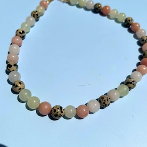 May include: A beaded necklace featuring a variety of colorful gemstones. The necklace includes round beads in shades of pink, green, white, and speckled brown. The necklace is strung on a gold-colored chain.