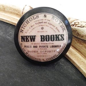 May include: A round, black decorative knob with the text "NEW BOOKS" in a vintage style. The knob features the text "NICHOLS & NOYES" and "BOSTON" in a circular design. The knob is likely for a drawer or cabinet.