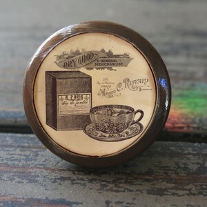 May include: A round wooden knob with a vintage design featuring a teacup and saucer, a box of tea, and the text "Dry Goods & General Merchandise" and "A Paris this de jardin".