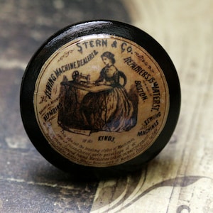 May include: A black wooden knob with a paper label featuring a woman sewing on a machine. The label reads "Stern & Co. Sewing Machine Dealers & Repairers, 15 Water St. Boston. Sewing Machines of all Kinds."