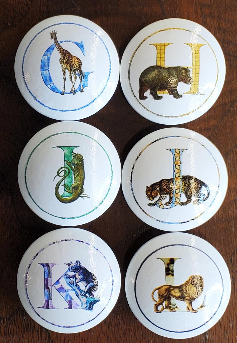May include: Six white ceramic cabinet knobs with animal illustrations and letters. The knobs feature a giraffe, bear, iguana, leopard, koala, and lion. Each animal is paired with a letter of the alphabet.