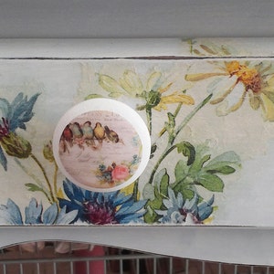 May include: A round white drawer knob featuring a printed image of small birds perched on a branch, set against a light blue drawer front decorated with a decoupage floral pattern of blue cornflowers and yellow daisies with green leaves.