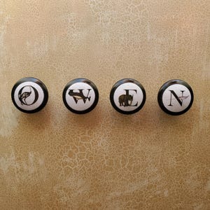 以下が含まれることがあります： 黒い縁と白い中心部を持つ4つの装飾的なキャビネットノブ。各ノブには文字と動物のイラストが描かれています。ダチョウ、クジラ、ゾウ、鳥です。ノブは、テクスチャ加工されたベージュ色の背景に並んで配置されています。