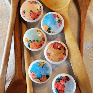 May include: Six floral patterned ceramic knobs with a variety of colors and designs, including pink, blue, yellow, and green, are arranged on a white surface with four wooden spoons.