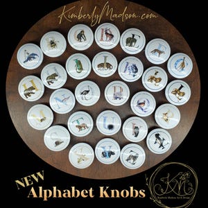 May include: A collection of white alphabet knobs with animal illustrations and letters. The knobs are arranged on a dark wooden surface. The text "NEW Alphabet Knobs" is visible at the bottom of the image.