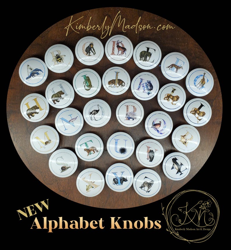 May include: A selection of white alphabet knobs featuring animal designs and letters. The knobs are displayed on a dark wooden surface. The text "NEW Alphabet Knobs" is visible at the bottom of the image.