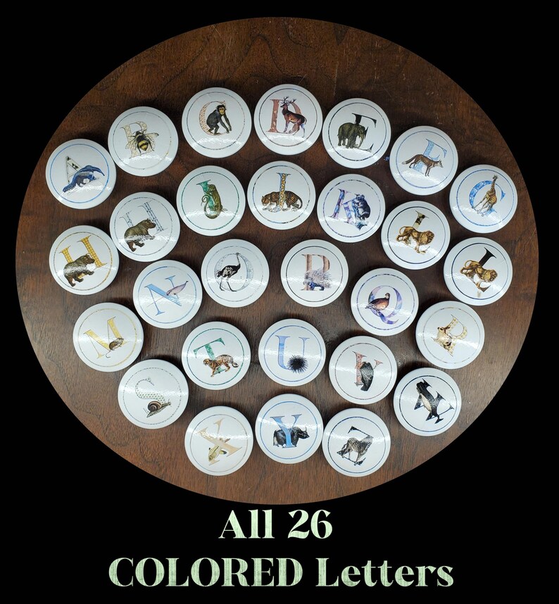 May include: A set of 26 decorative knobs, each with a letter of the alphabet and an animal illustration. The knobs are white and round, arranged on a dark wooden surface. The text at the bottom reads "All 26 COLORED Letters."