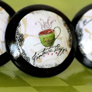May include: Three black cabinet knobs with coffee-themed designs. One knob features a green coffee cup with the words "Fantastic Caffe". The other knobs have coffee cup illustrations and decorative elements.