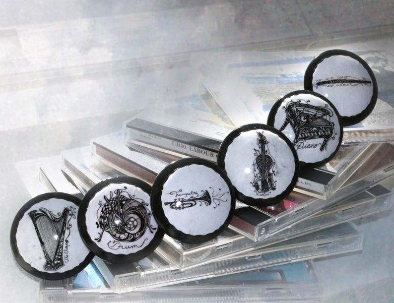 May include: Six round black and white coasters with musical instruments drawn on them. The coasters are arranged in a row on top of a stack of CDs. The instruments include a harp, a drum, a trumpet, a violin, a piano, and a flute. The coasters are all white with black outlines and the instruments are drawn in black ink. The CDs are all silver with black labels. The text on the coasters reads: 'Harp', 'Drum', 'Trumpet', 'Violin', 'Piano', and 'Flute'.