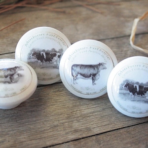 May include: Four white ceramic knobs with black and white cow illustrations and the text "Madison Farms Cattle Co. Angus Beef" printed on each knob.