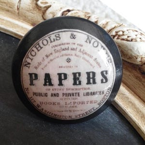 May include: A round, black object with a vintage label featuring the text "PAPERS" in bold letters. The label also includes the text "NICHOLS & NO" and "BOOKS IMPORTED". The object is set against a dark surface and a decorative frame.