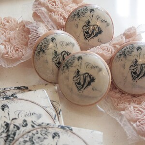 May include: Four decorative knobs with a white background and black floral designs. Each knob features a woman in a dress and the word "Ciao" written on the knob. The knobs are surrounded by pink fabric with a floral design.