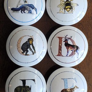 May include: A collection of six white ceramic cabinet knobs, each displaying a letter and an animal illustration. The letters are A, B, C, D, E, and F. The animals depicted are an anteater, bee, monkey, deer, elephant, and fox.