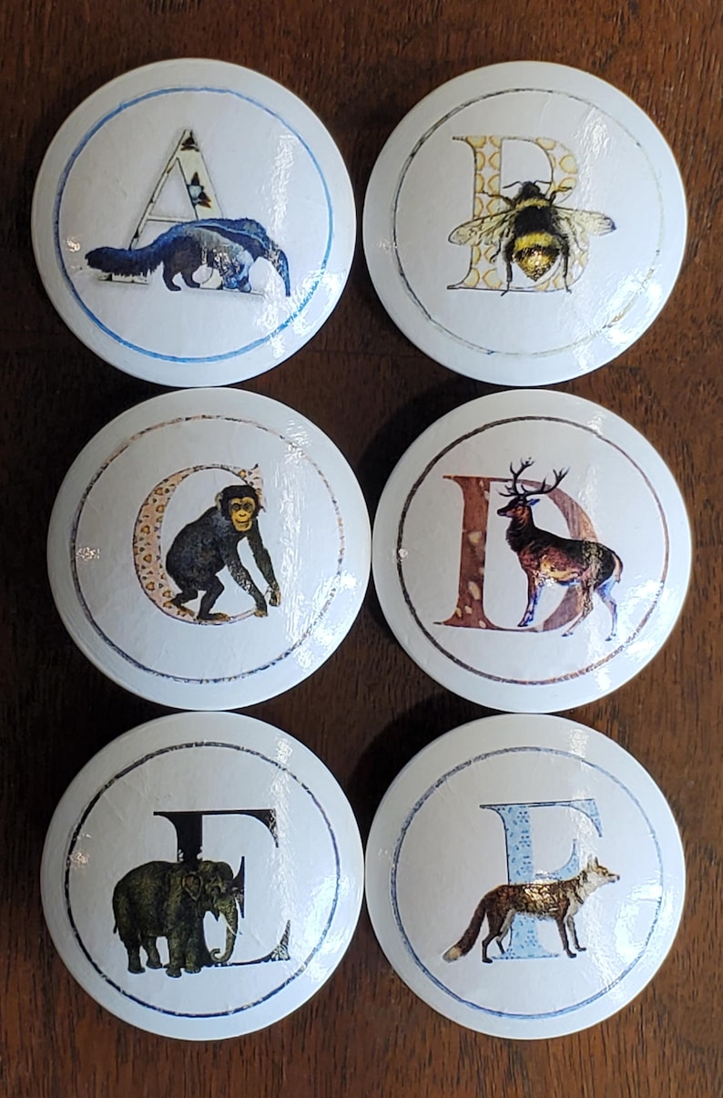 May include: A collection of six white ceramic cabinet knobs, each displaying a letter and an animal illustration. The letters are A, B, C, D, E, and F. The animals depicted are an anteater, bee, monkey, deer, elephant, and fox.