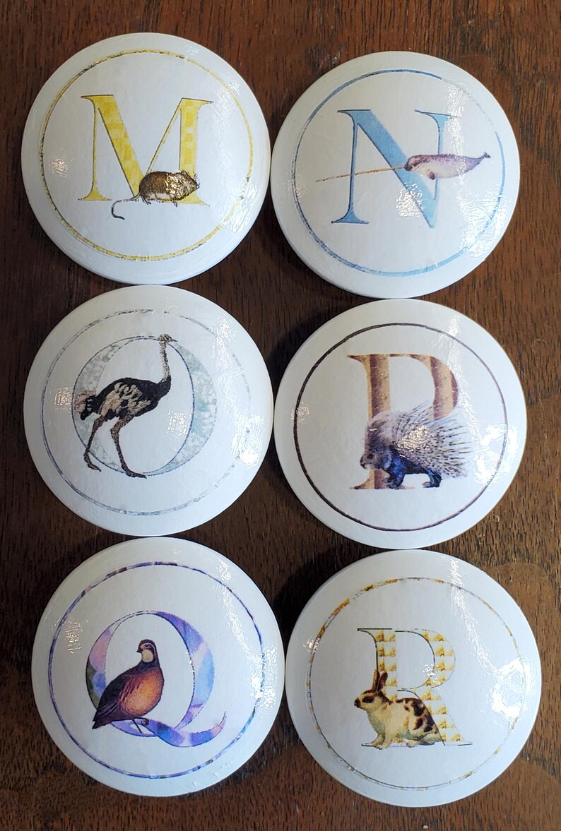 May include: Six white cabinet knobs with letters and animal illustrations. The knobs feature the letters M, N, O, P, Q, and R, each paired with a corresponding animal: a mouse, narwhal, ostrich, porcupine, quail, and rabbit.