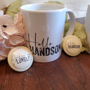 May include: White ceramic mug with black text that reads "Hello Handsome".  Two round, beige coasters with black text. One coaster reads "Good morning Lovely" and the other reads "Hello Handsome".