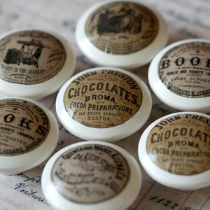 May include: A collection of seven white ceramic cabinet knobs with vintage-style labels. The labels feature text and illustrations in sepia tones, including the words "BOOKS" and "CHOCOLATES".
