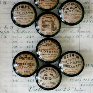 May include: Nine vintage black and white round knob-like objects with various advertisements printed on them. The advertisements include: "J.W. Black Photographs", "Geo. H. Richards Men's & Boys Clothing", "Geo. N. Hood Demigant", "Jackman & Merril Clothing", "The Greatorgan in the Boston Music Hall", "Stumcke & Goodwine Dining Rooms for Ladies & Gent", "H.G. Smith Photograper & Publisher of Works of Art", and "Ahistons Dining Rooms for Ladies & Gentlemen".