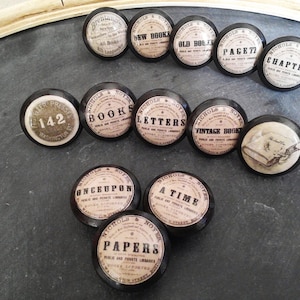 May include: A set of 11 black and white round knobs with various book-themed designs. The knobs feature text such as "New Books", "Old Books", "Vintage Books", "Letters", "Chapter", "Page 77", "Once Upon", "A Time", "Papers", and "Books".