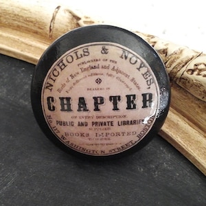 May include: A round, black decorative knob with a vintage label. The label reads "NICHOLS & NOYES" and "CHAPTER" in a classic font. The knob is likely for a drawer or cabinet, adding a touch of antique charm.