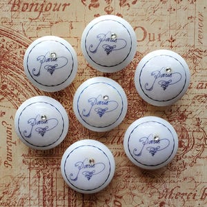 May include: White ceramic cabinet knobs with a blue script "Paris" design and a small crystal accent. The knobs are round and have a vintage aesthetic. The background has a sepia-toned, antique paper design.