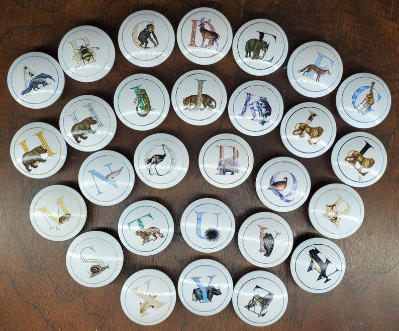May include: A selection of white cabinet knobs, each displaying a letter of the alphabet and an animal illustration. The circular knobs are arranged on a brown surface. The animal illustrations include a bee, bear, and zebra. Each knob is approximately 4 cm in diameter.