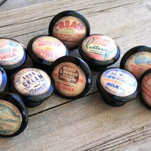 May include: A collection of 11 vintage-style milk bottle cap knobs with black bases. The knobs feature various milk brands and labels, including "Sour Cream", "Milk Raw", "Buttermilk", and "Pasteurized Milk".