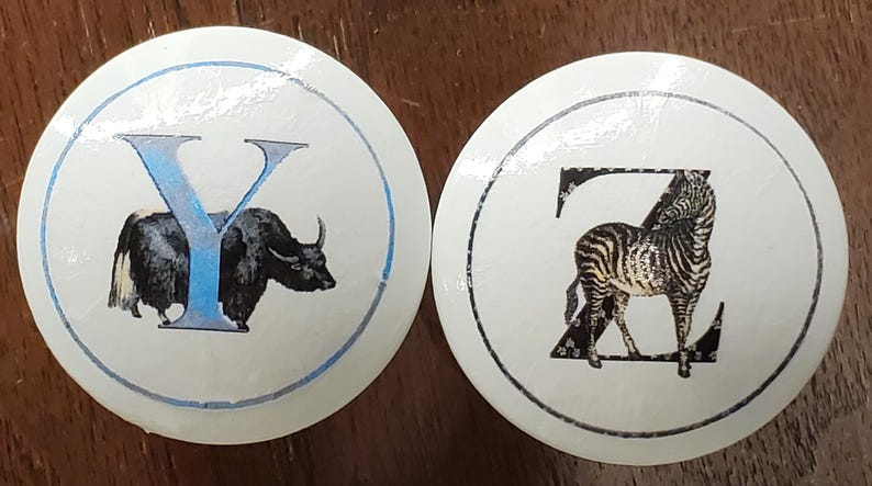 May include: Two white, round cabinet knobs. One features a blue "Y" with a yak illustration, the other a black "Z" with a zebra illustration. Both have a blue and black border.