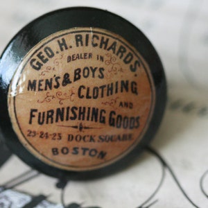 May include: A black, round knob with a vintage label. The label reads "GEO. H. RICHARDS DEALER IN MEN'S & BOYS CLOTHING AND FURNISHING GOODS" with the address "23-24-25 DOCK SQUARE BOSTON".