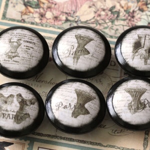 May include: Six black and white round knobs with a vintage Parisian theme. Each knob features a different image of a corset, a dress, or a hat with the word "Paris" printed on the knob.