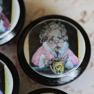 May include: A round black cabinet knob featuring a glossy circular insert with a watercolor illustration of an elderly person with curly gray hair and glasses wearing a pink coat, holding a yellow mug with a heart design and steam rising.
