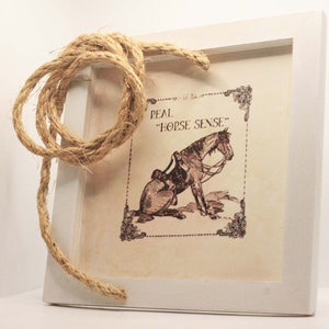 May include: A framed print with a sepia-toned illustration of a saddled horse, with the text "REAL HORSE SENSE." A thick, natural rope is looped over the white frame. The artwork has an antique, rustic aesthetic.