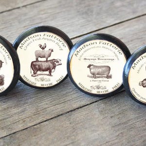 May include: Four black round knobs with white labels featuring vintage illustrations of farm animals and text that reads "Madson Fattorie". The labels include text in Italian and English.