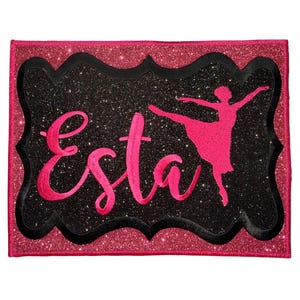 May include: A rectangular patch with a pink glitter border and a black glitter center. The patch features the name "Esta" in bright pink script lettering alongside a silhouette of a ballerina in a matching pink color, all with a black outline.