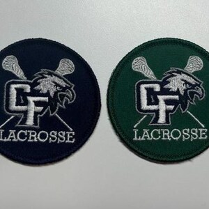 May include: Two round embroidered patches. One is navy blue, the other is dark green. Both feature a white lacrosse stick design, a stylized eagle head, and the word "LACROSSE" in white. The patches are likely for sports apparel.