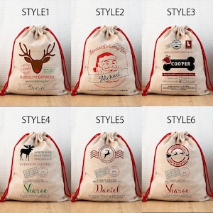 May include: A collection of six beige drawstring gift bags with red drawstrings. Each bag features a different holiday-themed design in red and brown, including reindeer, Santa Claus, and festive text. The bags are personalized with names.