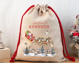 Può includere: Un sacco regalo natalizio in juta beige con coulisse rossa. La borsa presenta un'illustrazione festosa di Babbo Natale sulla sua slitta, renne e una scena di villaggio invernale. Il testo recita "Special Delivery For KENNETH".