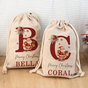 May include: Two beige drawstring gift bags with red lettering and Santa Claus illustrations. One bag has the letter "B" and the name "BELLA", the other has the letter "C" and the name "CORAL". Both bags say "Merry Christmas".