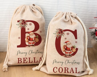 May include: Two beige drawstring gift bags with red lettering and Santa Claus illustrations. One bag has the letter "B" and the name "BELLA", the other has the letter "C" and the name "CORAL". Both bags say "Merry Christmas".