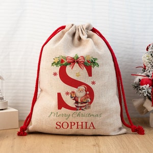 Pode incluir: Uma sacola de presente de Natal bege com fecho de cordão vermelho. A sacola apresenta uma grande letra vermelha "S", uma ilustração do Papai Noel e as palavras "Merry Christmas SOPHIA". Decorada com azevinho e flocos de neve, é festiva.