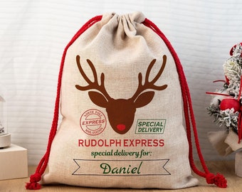 Può includere: Un sacco regalo natalizio beige con coulisse rossa. La borsa presenta una grafica a testa di renna marrone e il testo "RUDOLPH EXPRESS" e "special delivery for: Daniel". La borsa ha un timbro "North Pole Express Mail Station".