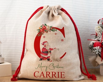 Può includere: Un sacco regalo natalizio beige con chiusura a coulisse rossa. La borsa presenta una grande lettera "C" rossa con un'illustrazione di Babbo Natale e le parole "Merry Christmas CARRIE" in verde e rosso.