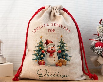 Può includere: Un sacco regalo natalizio beige con coulisse rossa. La borsa presenta un pupazzo di neve, alberi di Natale e scatole regalo. Il testo sulla borsa recita "SPECIAL DELIVERY FOR" e un nome. Design festivo natalizio.